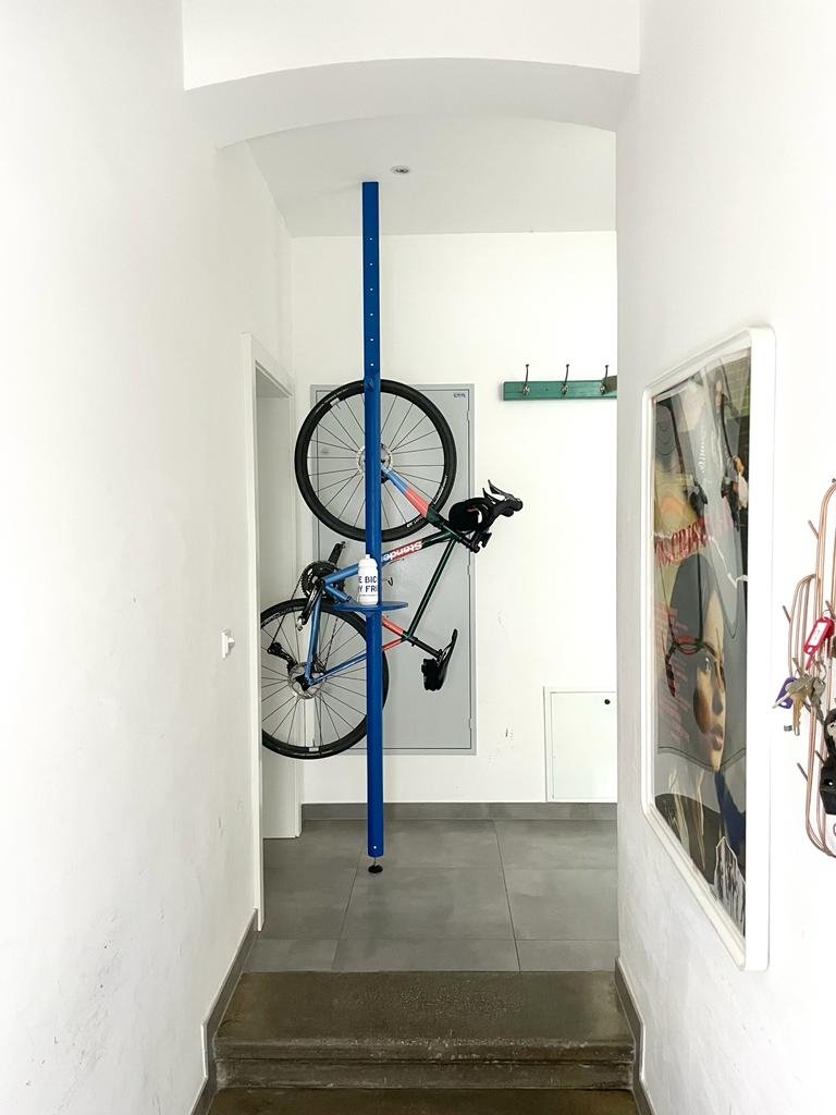 Vertical blue bike storage rack holding a black and gray road bicycle vertically against a white wall in a narrow hallway.