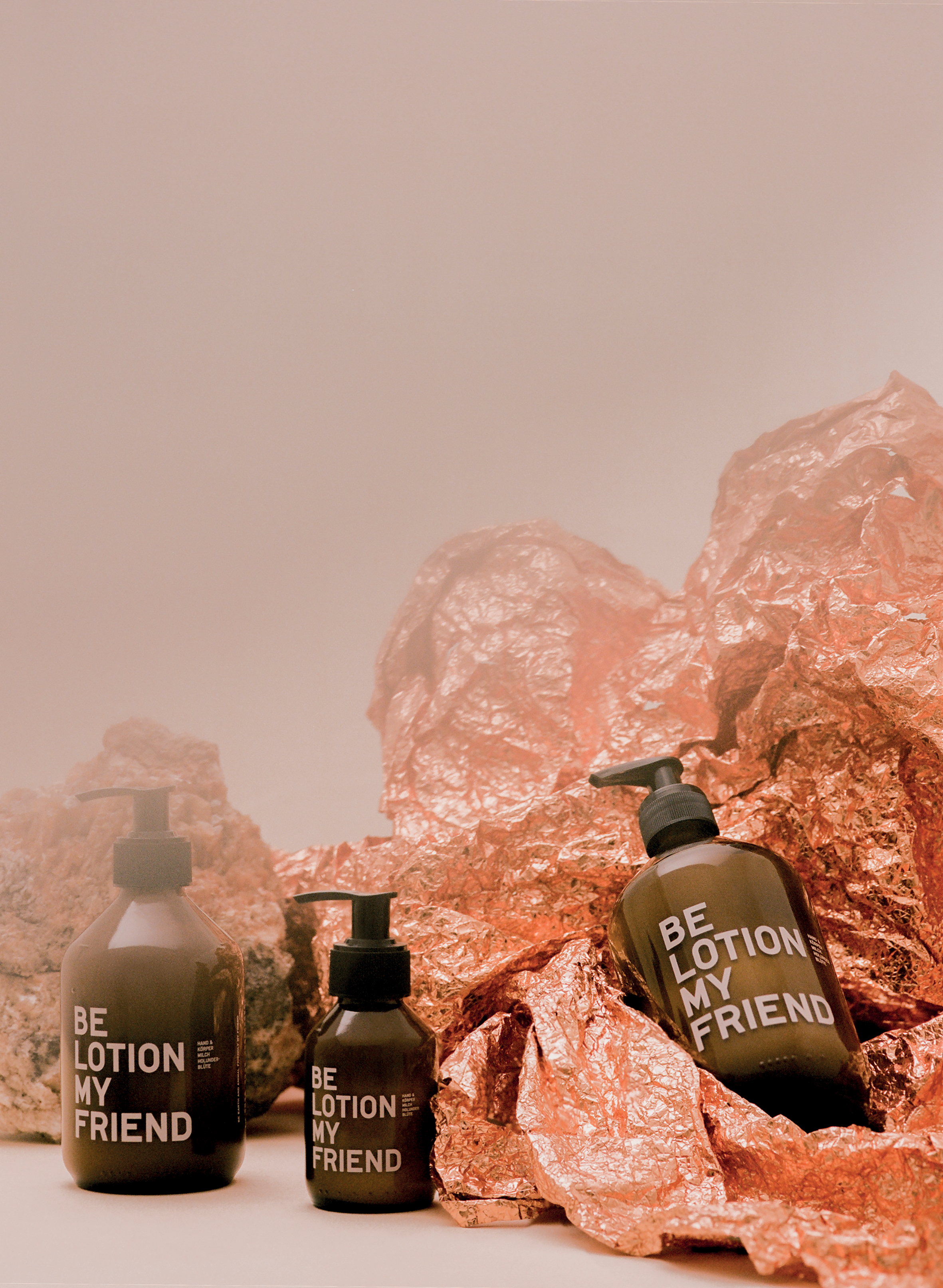 Two dark amber pump bottles labeled "Be Lotion My Friend" on crinkled rose gold foil against a soft beige background.