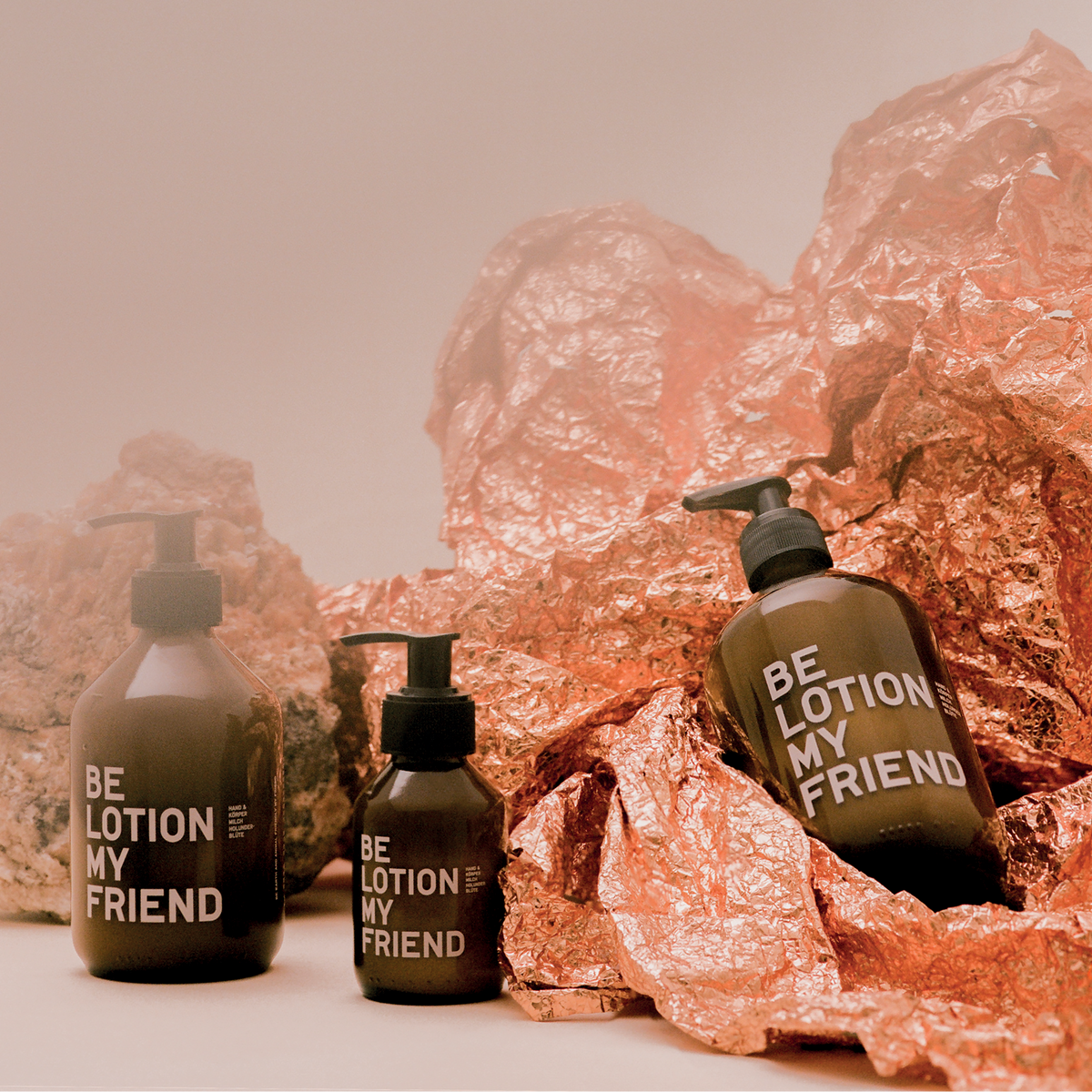 Two dark amber pump bottles labeled "Be Lotion My Friend" on crinkled rose gold foil against a soft beige background.