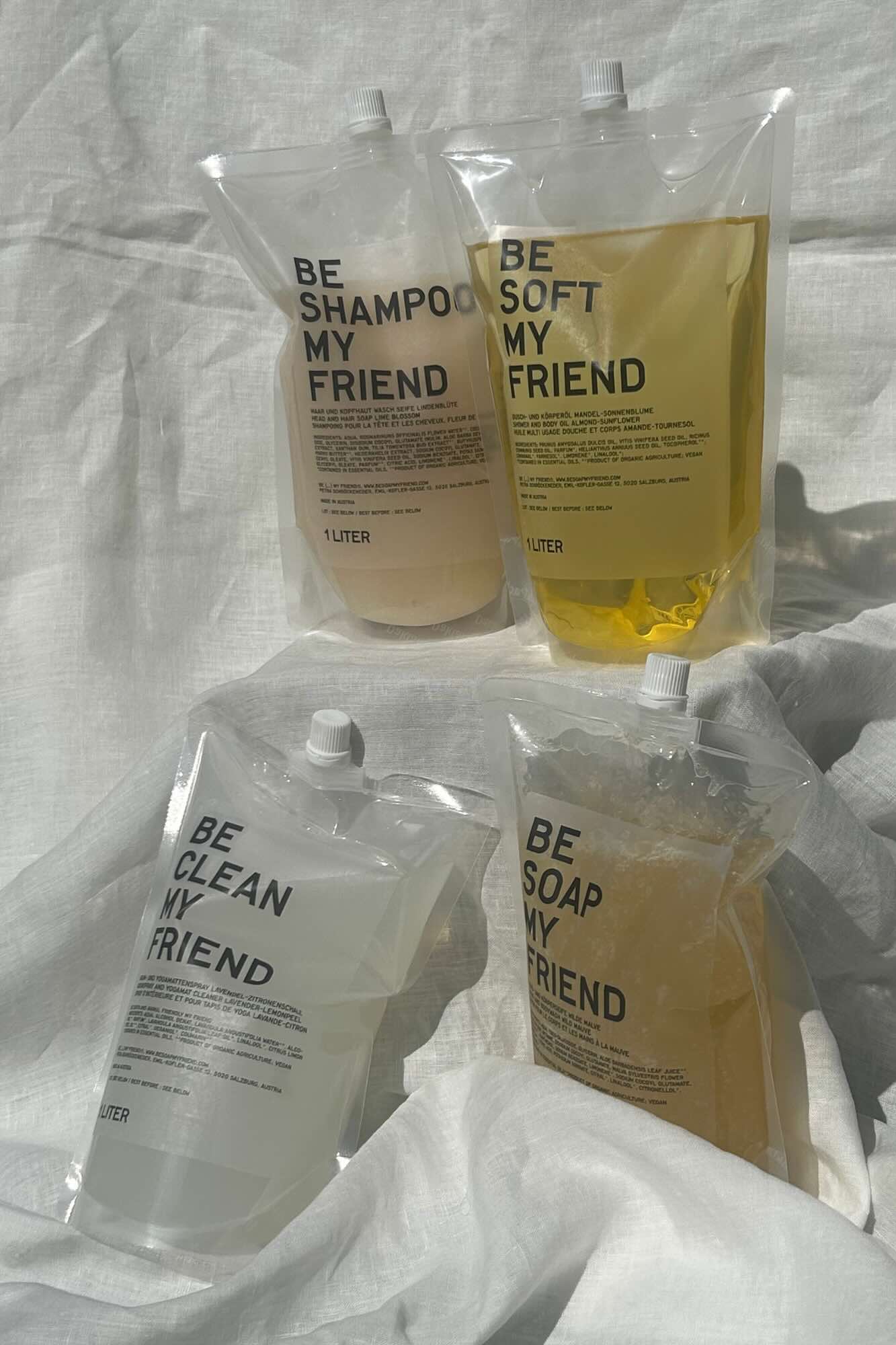 Four clear refill pouches labeled shampoo, body oil, cleanser, and soap with minimalist black text on a white cloth.