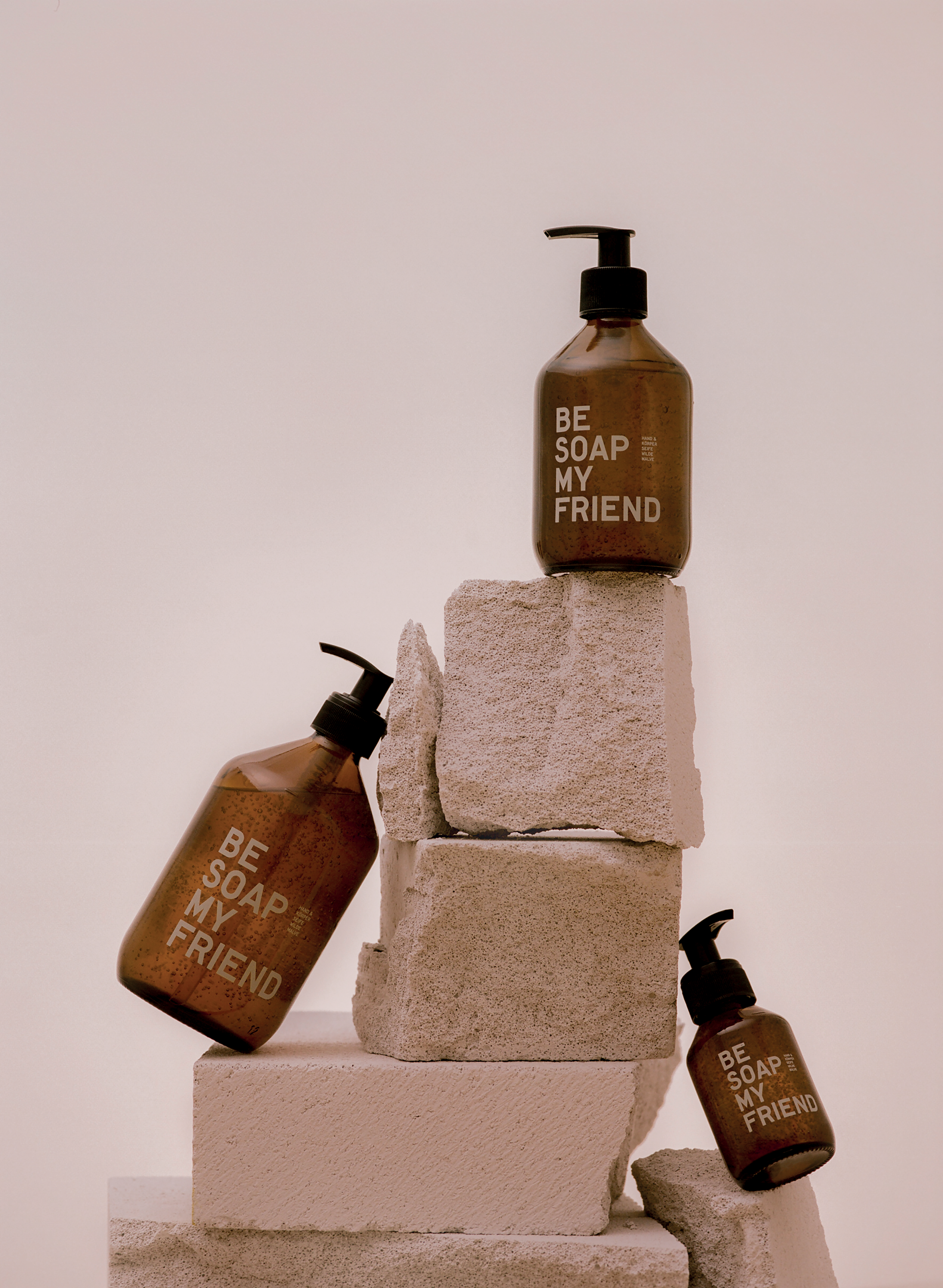 Set of three amber soap dispensers with black pumps, labeled "Be Soap Your Friend," stacked on beige stone blocks against a.