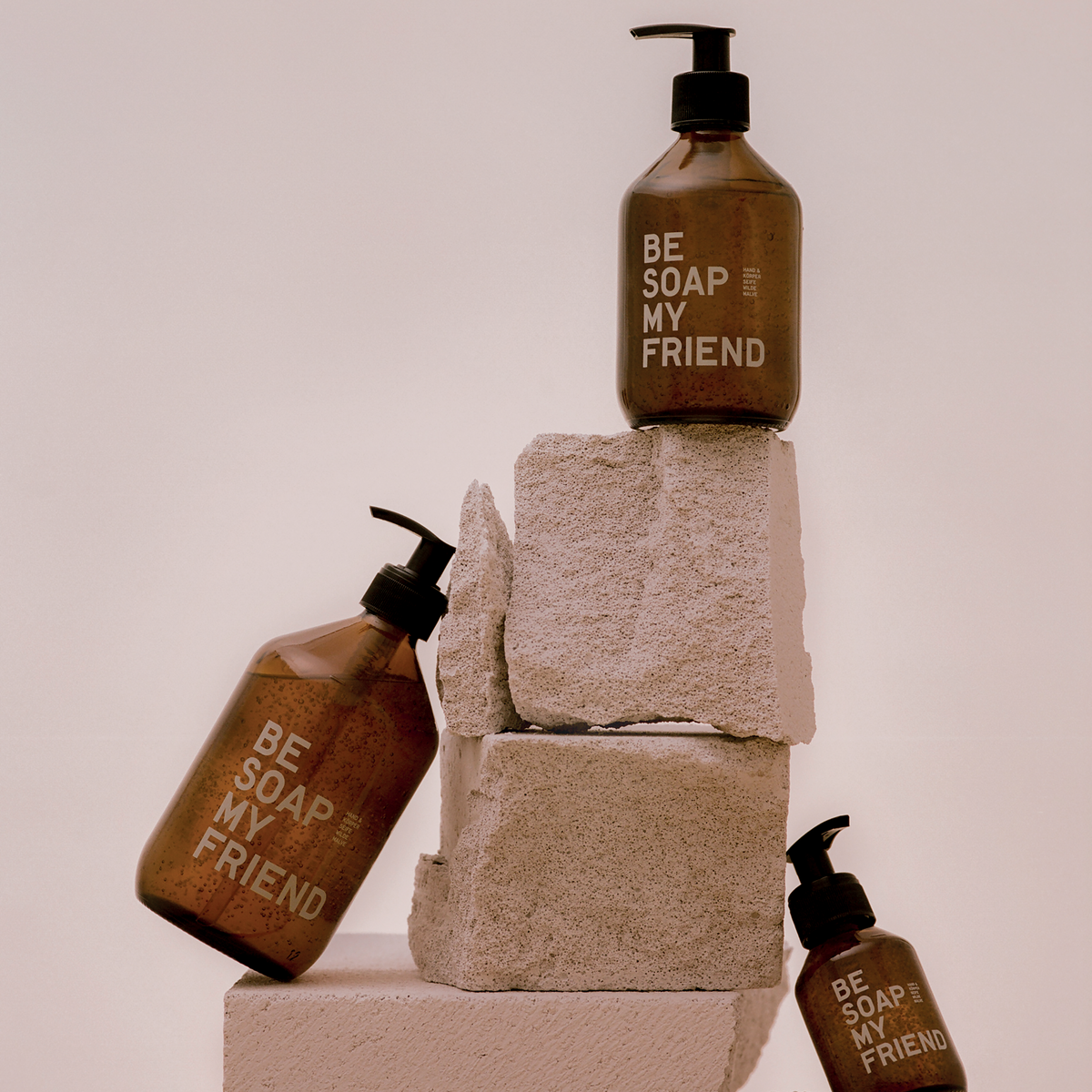 Set of three amber soap dispensers with black pumps, labeled "Be Soap Your Friend," stacked on beige stone blocks against a.