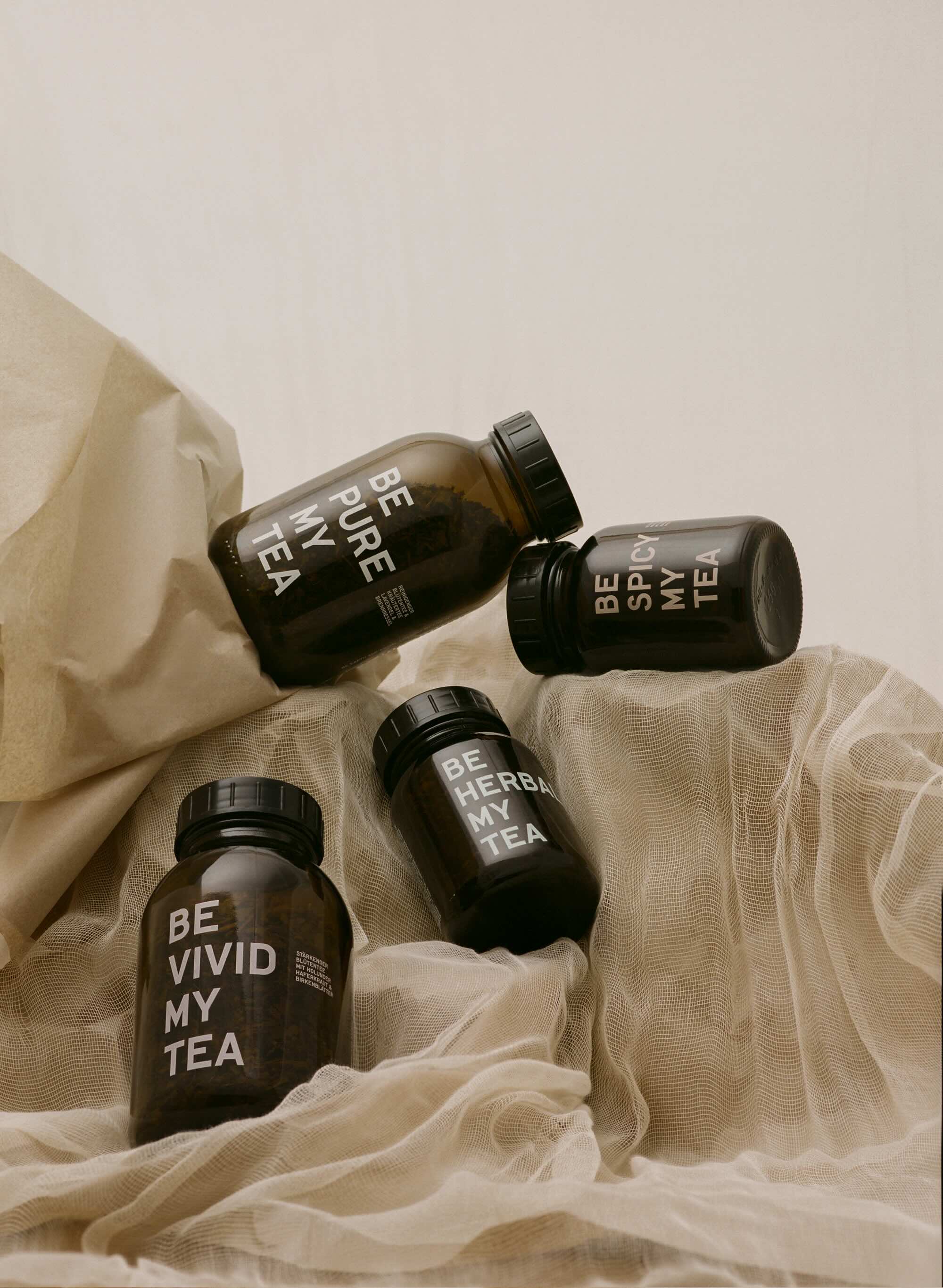 Four dark brown glass jars with black lids labeled "BE VIVID MY TEA" on light beige fabric background.