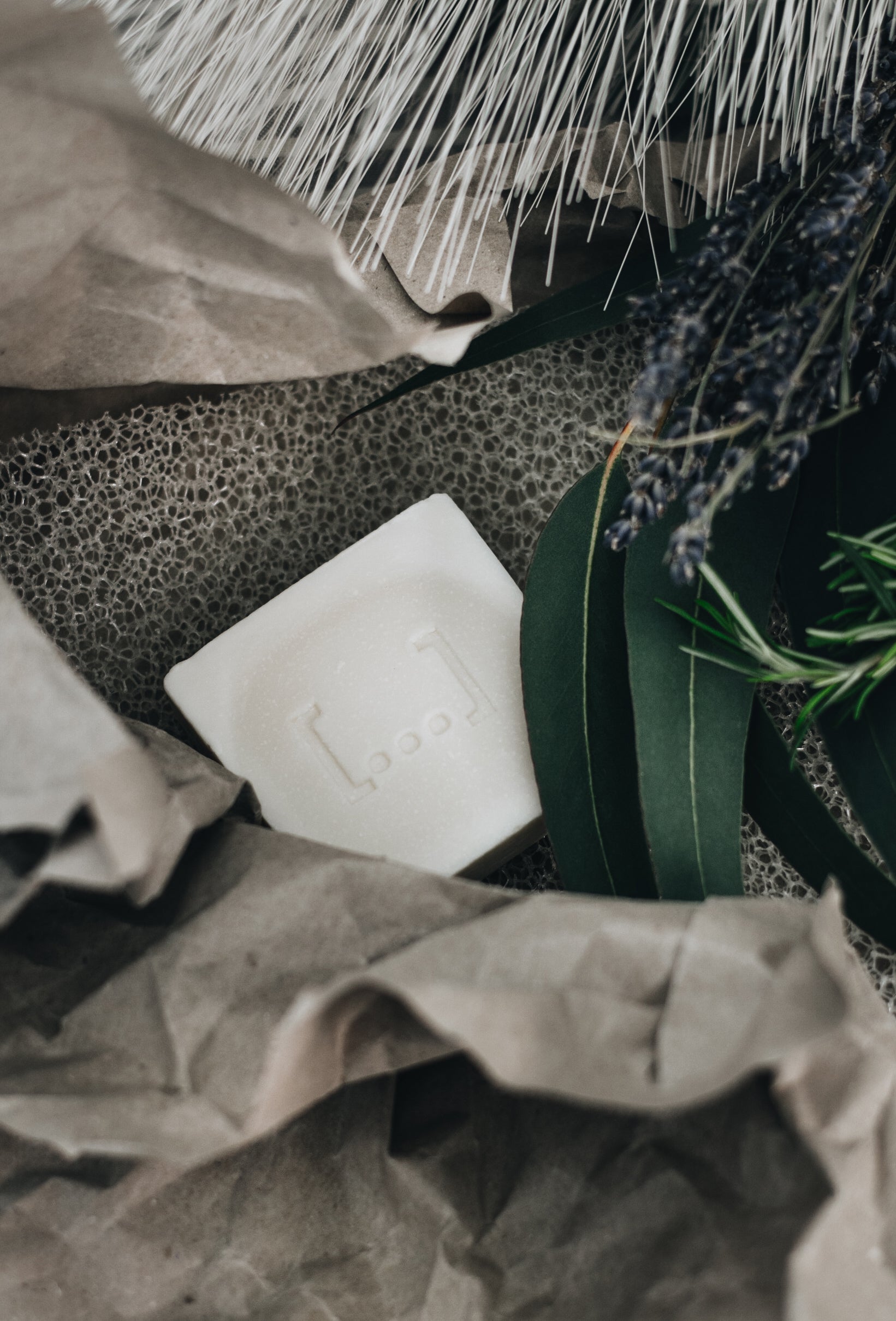 White square lavender soap bar with embossed logo, surrounded by brown paper and lavender sprigs on textured surface.