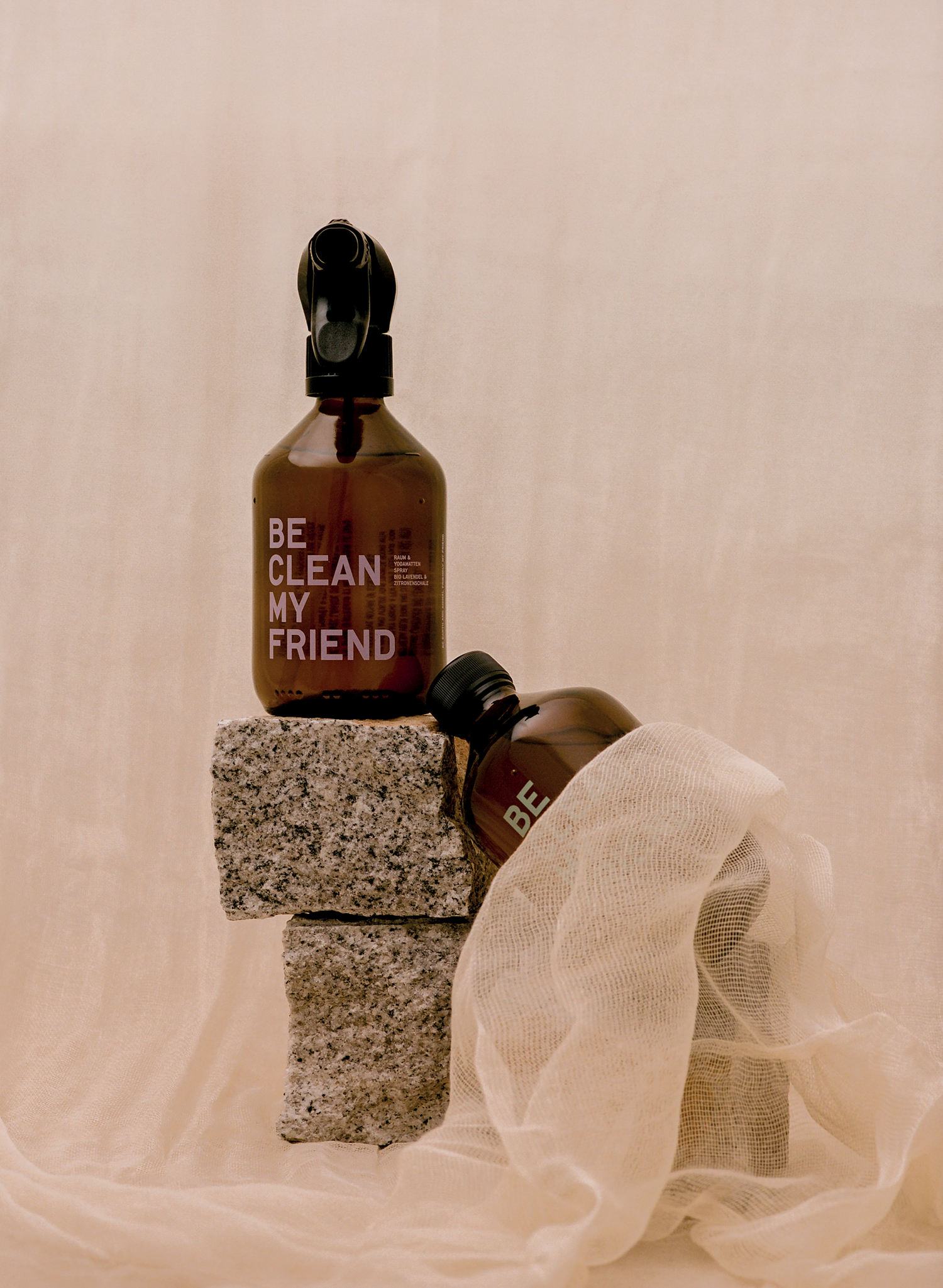 Amber glass spray bottles with black nozzles labeled "Be clean my friend" resting on textured gray blocks and soft beige.