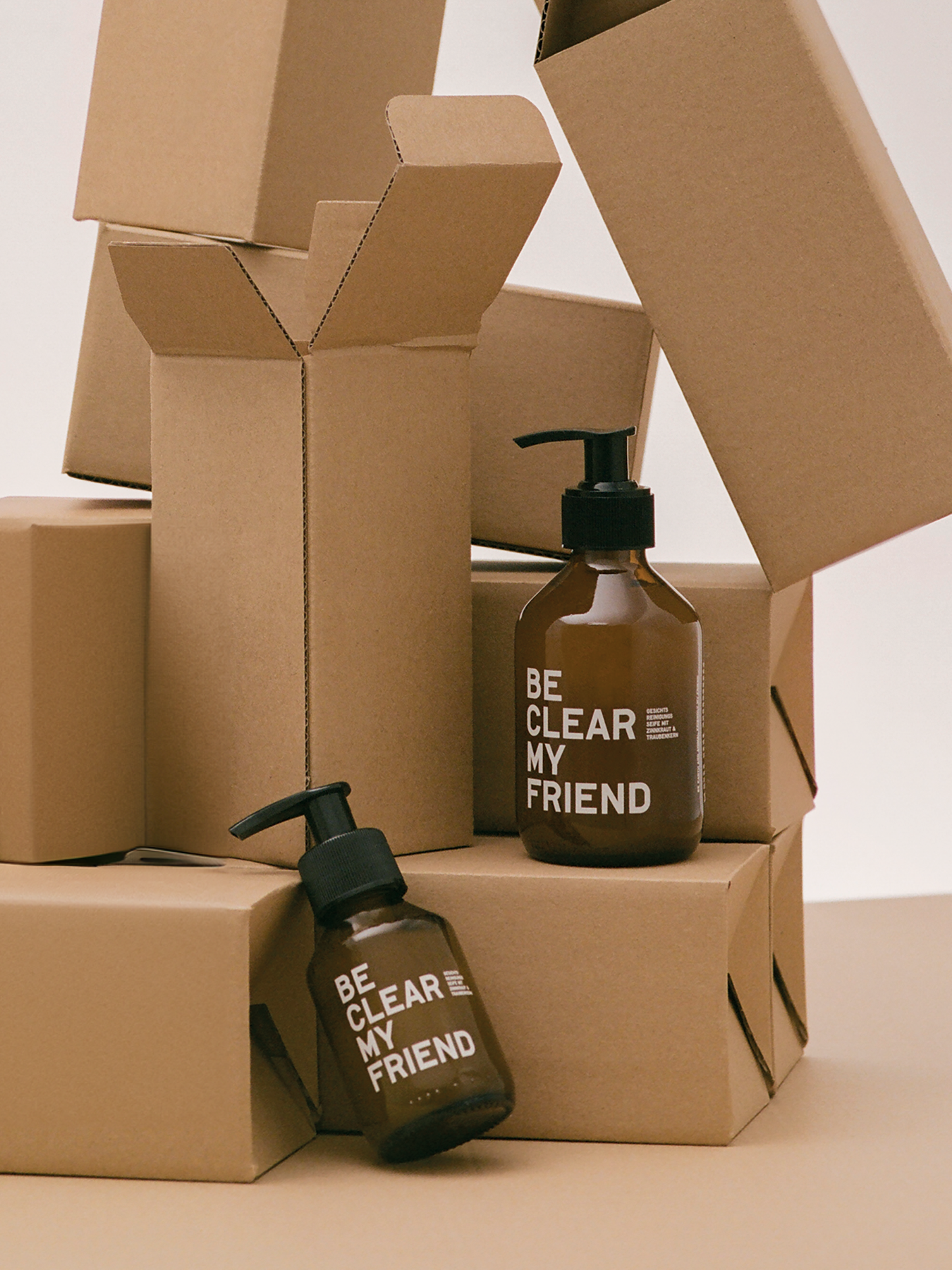 Brown pump bottle labeled "Be Clear My Friend" surrounded by various open and closed cardboard boxes on a beige surface.