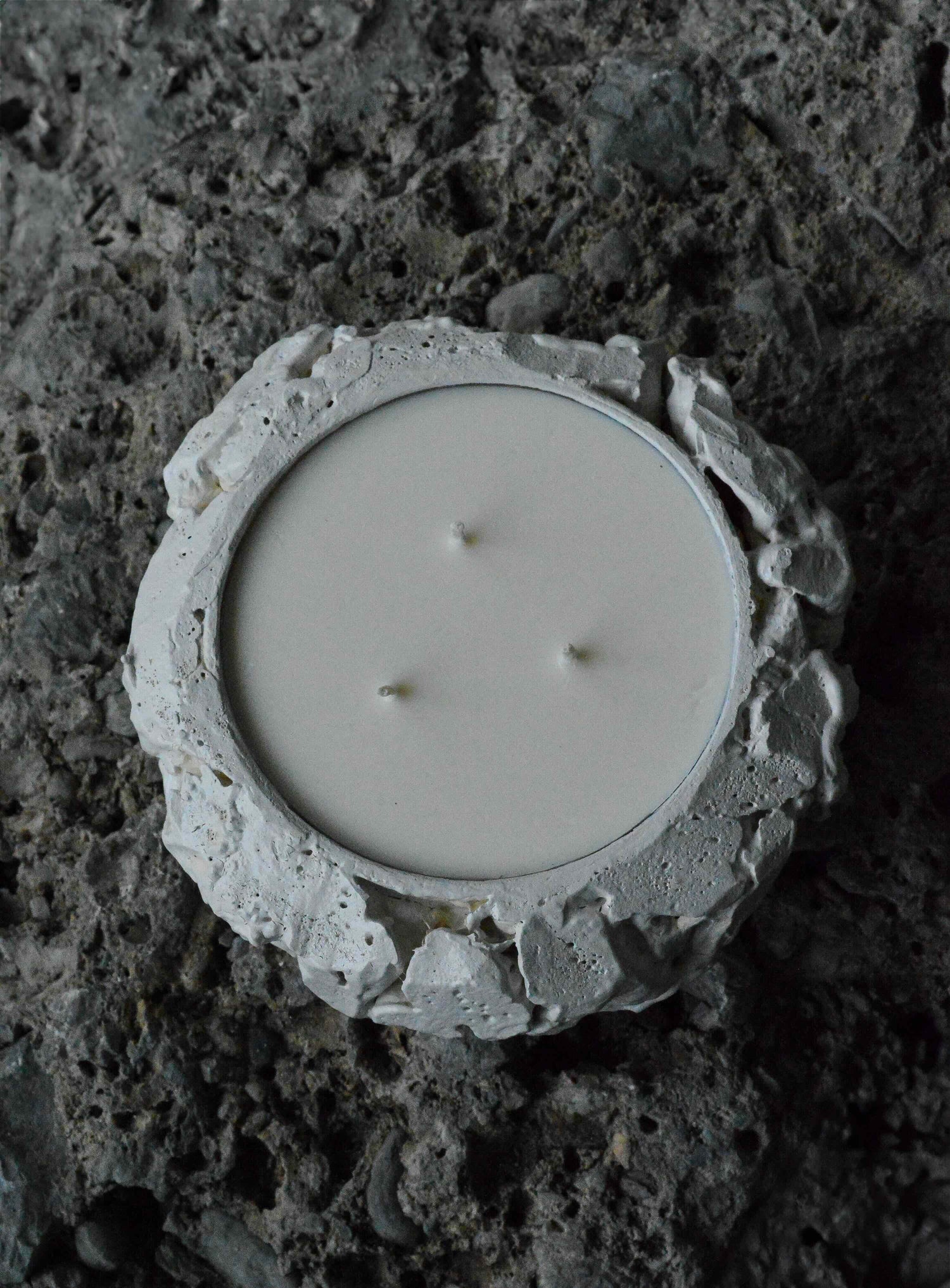 White three-wick candle in textured, rough-edged stone holder on dark stone surface.