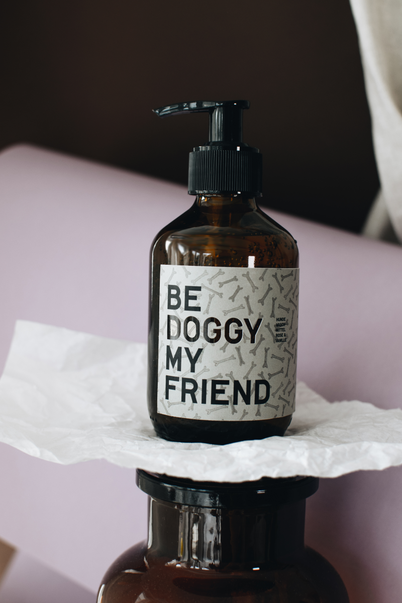 Amber pump bottle labeled "Be Doggy My Friend" on a crumpled white paper with soft pink and beige background.
