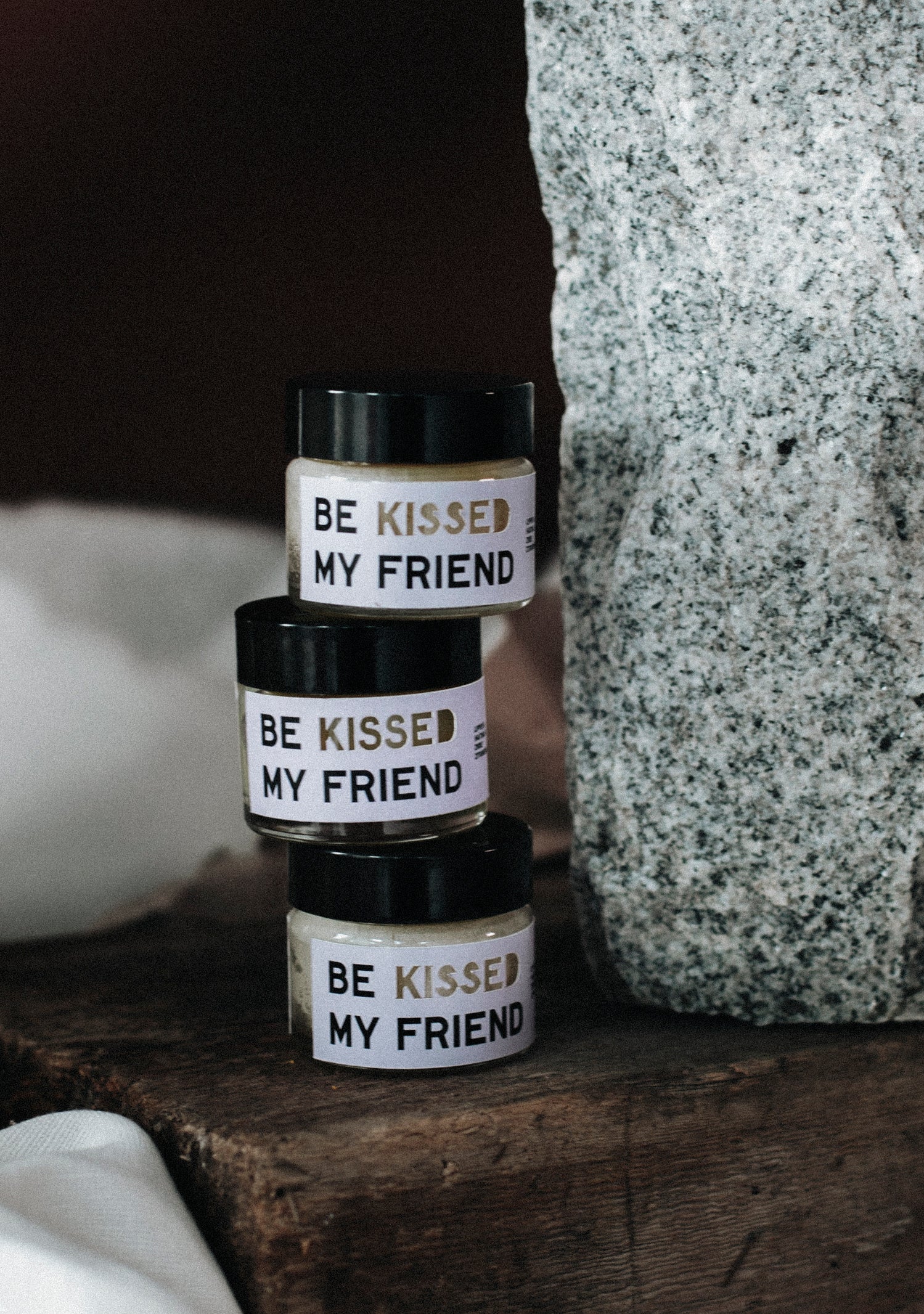 Three stacked jars of BE KISSED MY FRIEND lip and face balm with black lids and white labels, beside a stone textured surface.
