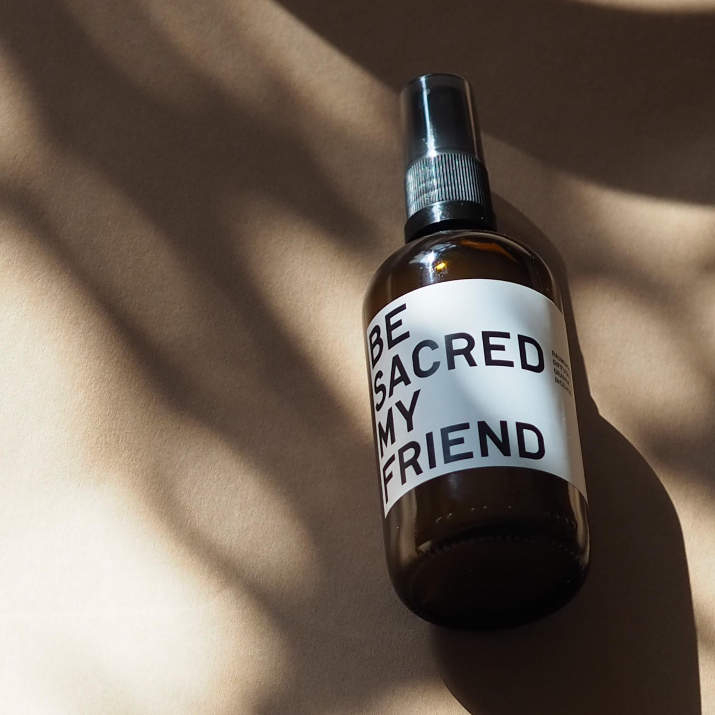Amber glass spray bottle with white label reading "BE SACRED MY FRIEND" on sunlit surface.
