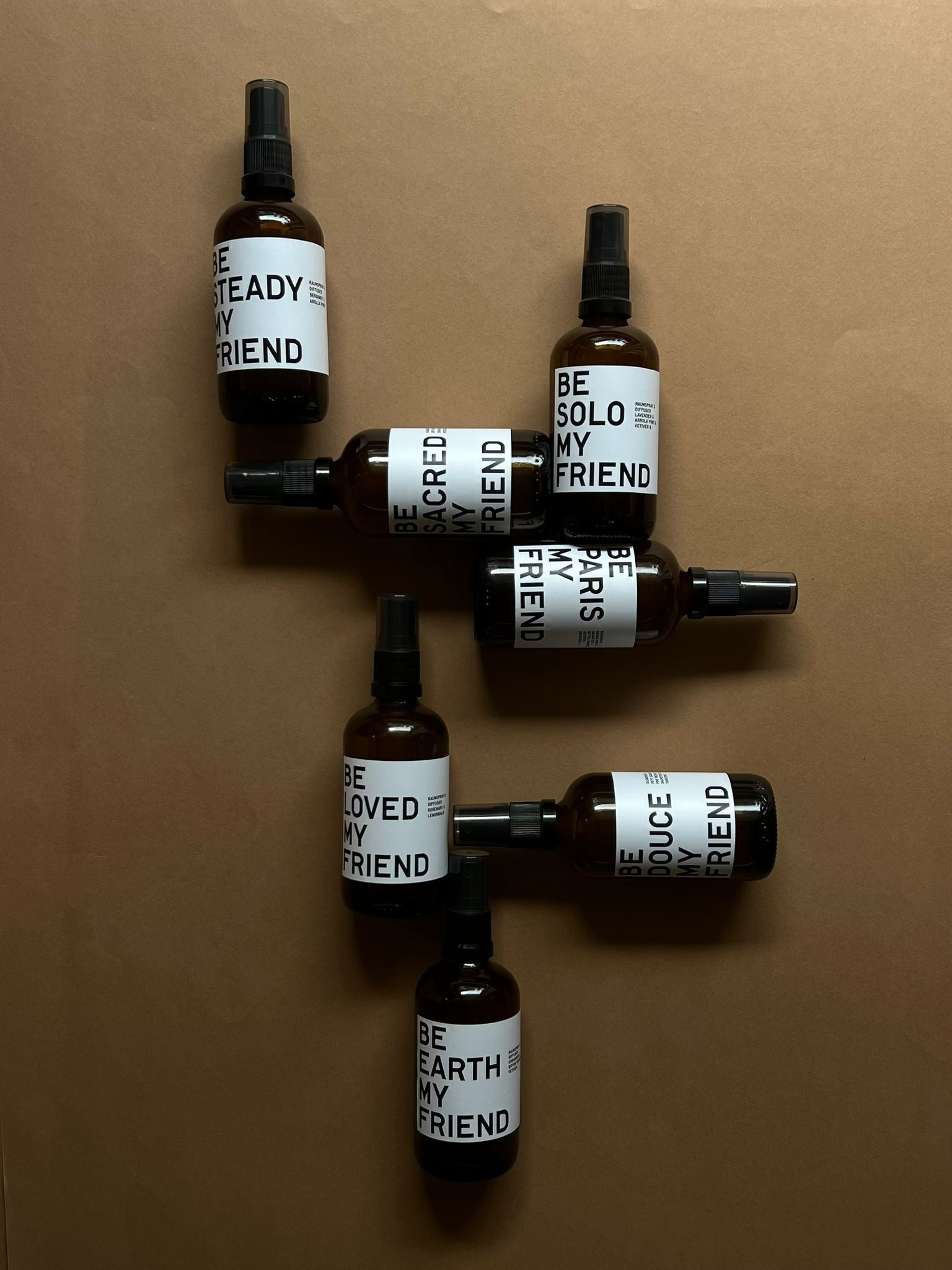 Six dark amber spray bottles with minimalist white labels and black text arranged on brown surface.