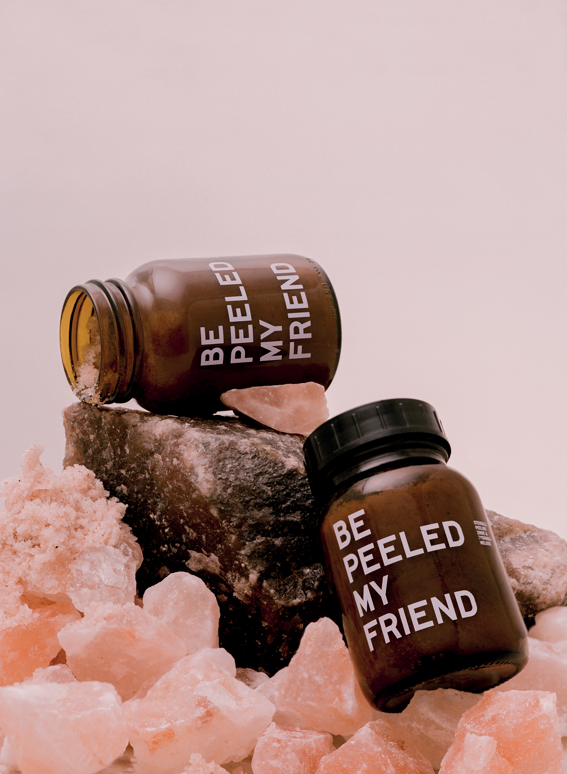 Two amber glass jars with black lids labeled "Be Peeled My Friend" and "Be Melt My Friend" on pink salt crystals and a dark.