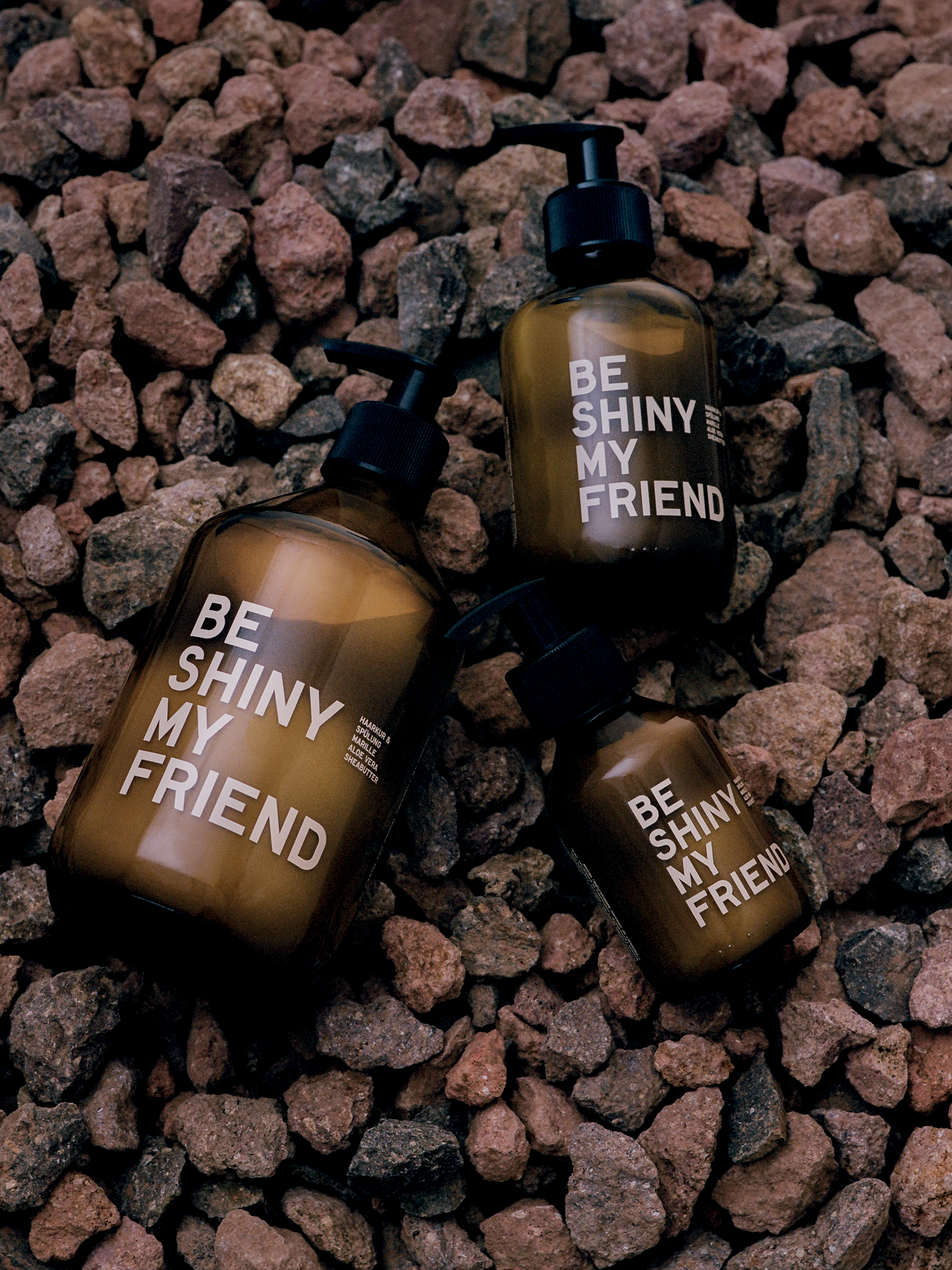 Three amber pump bottles labeled "Be Shiny My Friend" on brown gravel background.