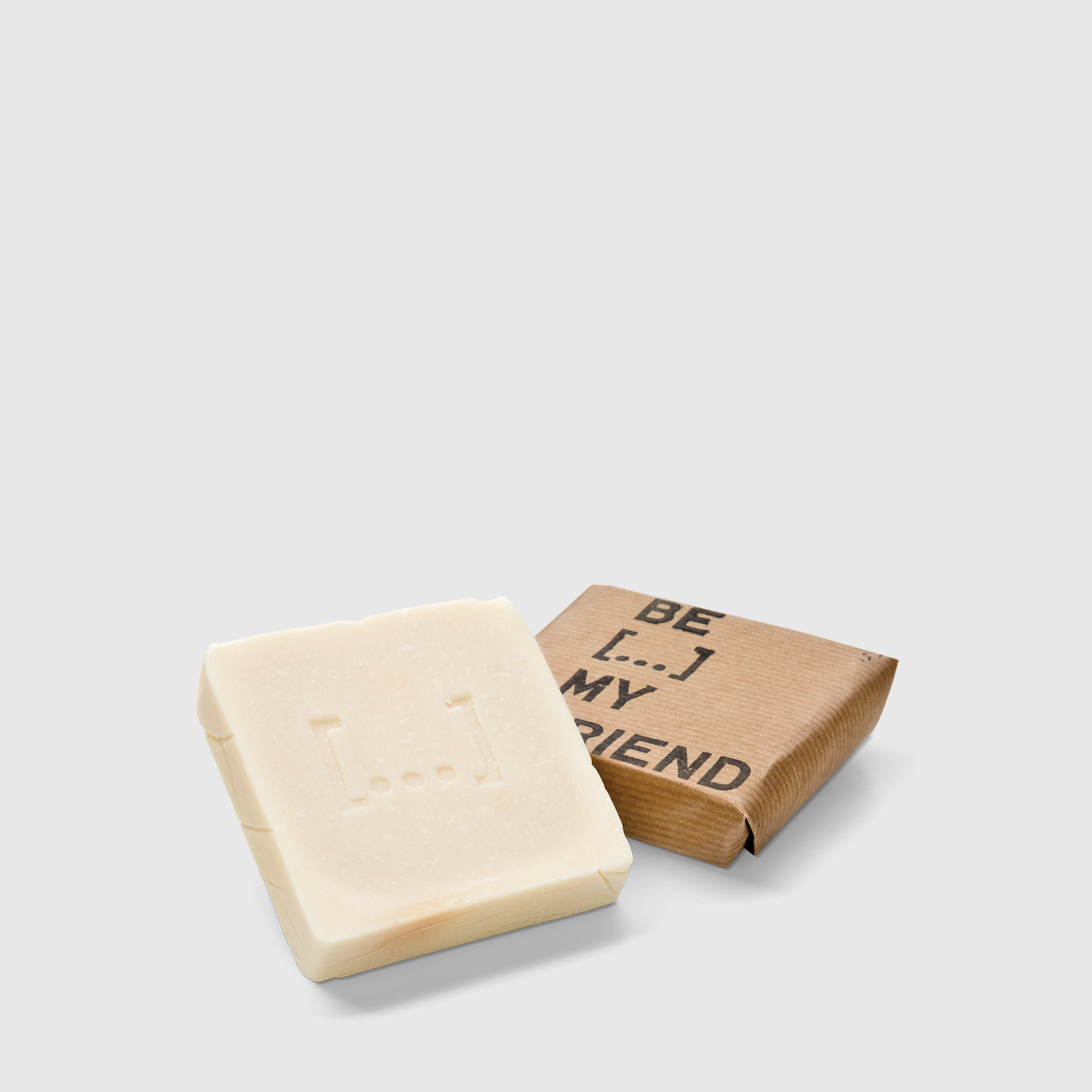 Creamy white square soap bar with embossed logo next to brown paper packaging labeled "Be My Friend".