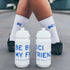 White cycling socks with blue "BE BICI" text worn with black shoes, white water bottles with blue text in foreground.