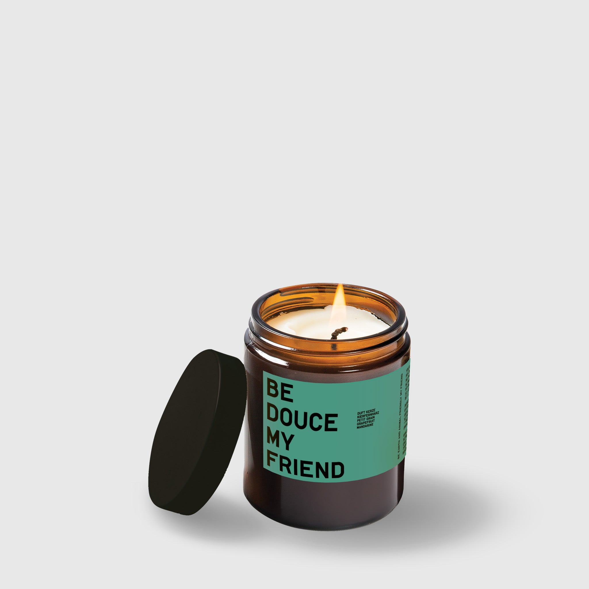 Amber glass candle jar with mint green label reading "Be Dque My Friend" and black lid beside it.