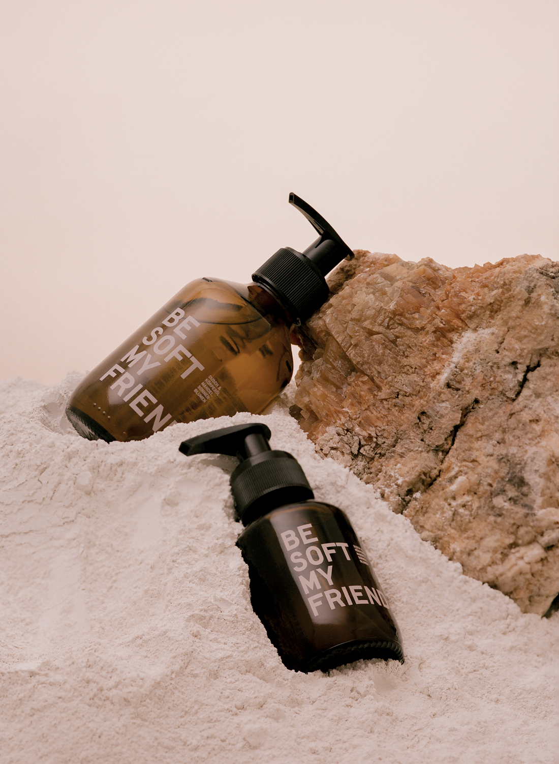 Two brown pump bottles labeled "Be Soft My Friend" placed on white sand next to a rough beige stone.