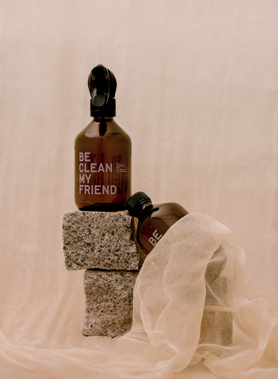 Amber glass spray bottles with black nozzles labeled "Be clean my friend" resting on textured gray blocks and soft beige.