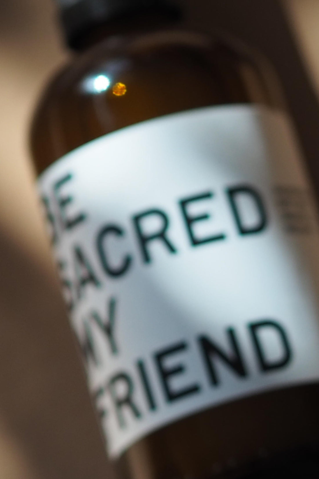 Amber glass spray bottle with white label reading BE SACRED INTERIOR FRIEND in black text.