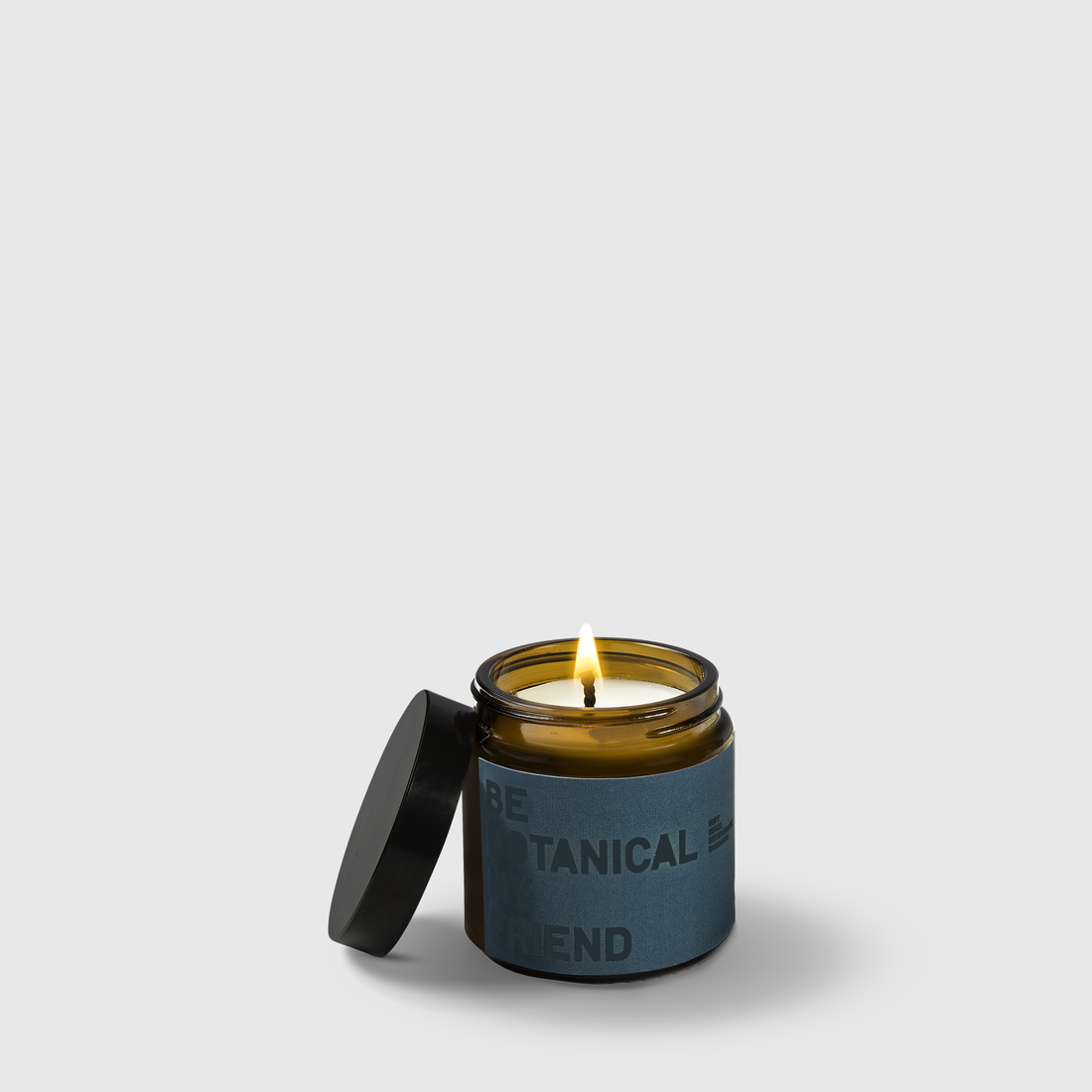 Amber glass candle jar with burning wick and dark blue label reading Botanical Blend, black lid beside jar.