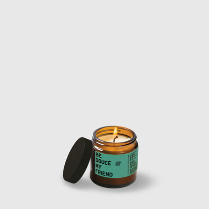Amber glass candle jar with teal label reading "Be Douce My Friend" and black lid beside it.