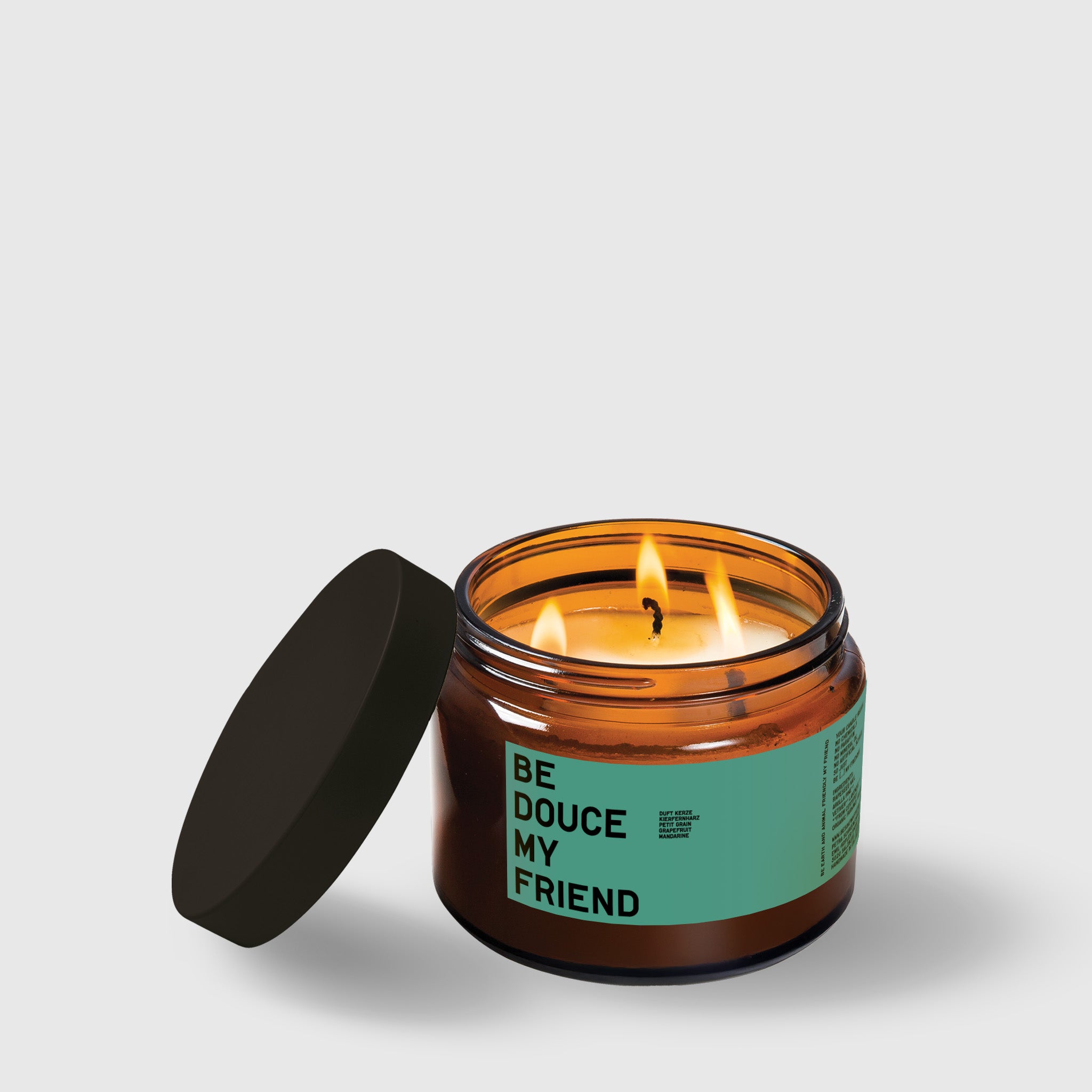 Amber glass candle jar with black lid and "Be Loved My Friend" label, lit wick visible.