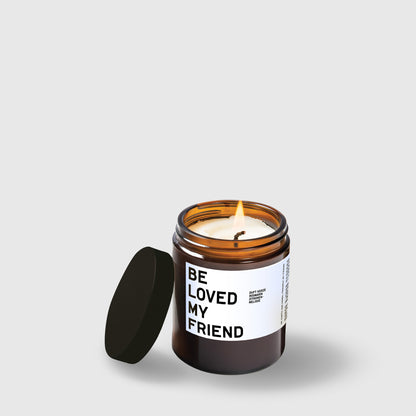 Amber glass candle jar with black lid and "Be Loved My Friend" text label, lit wick visible.