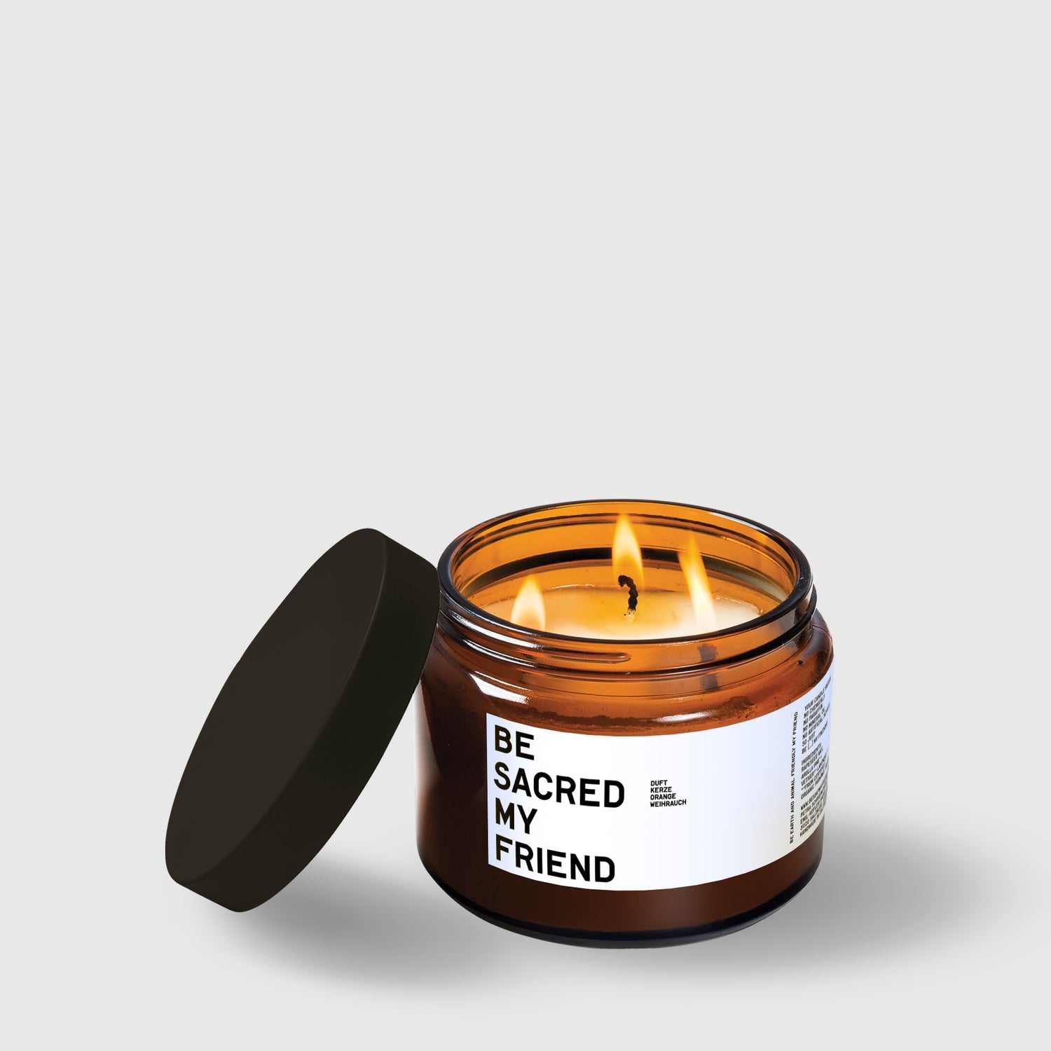 Amber glass candle jar labeled "Be Sacred My Friend" with black lid and lit wick.
