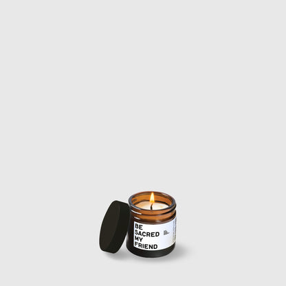 Lit amber glass candle with black lid and white label reading "Be sacred my friend".